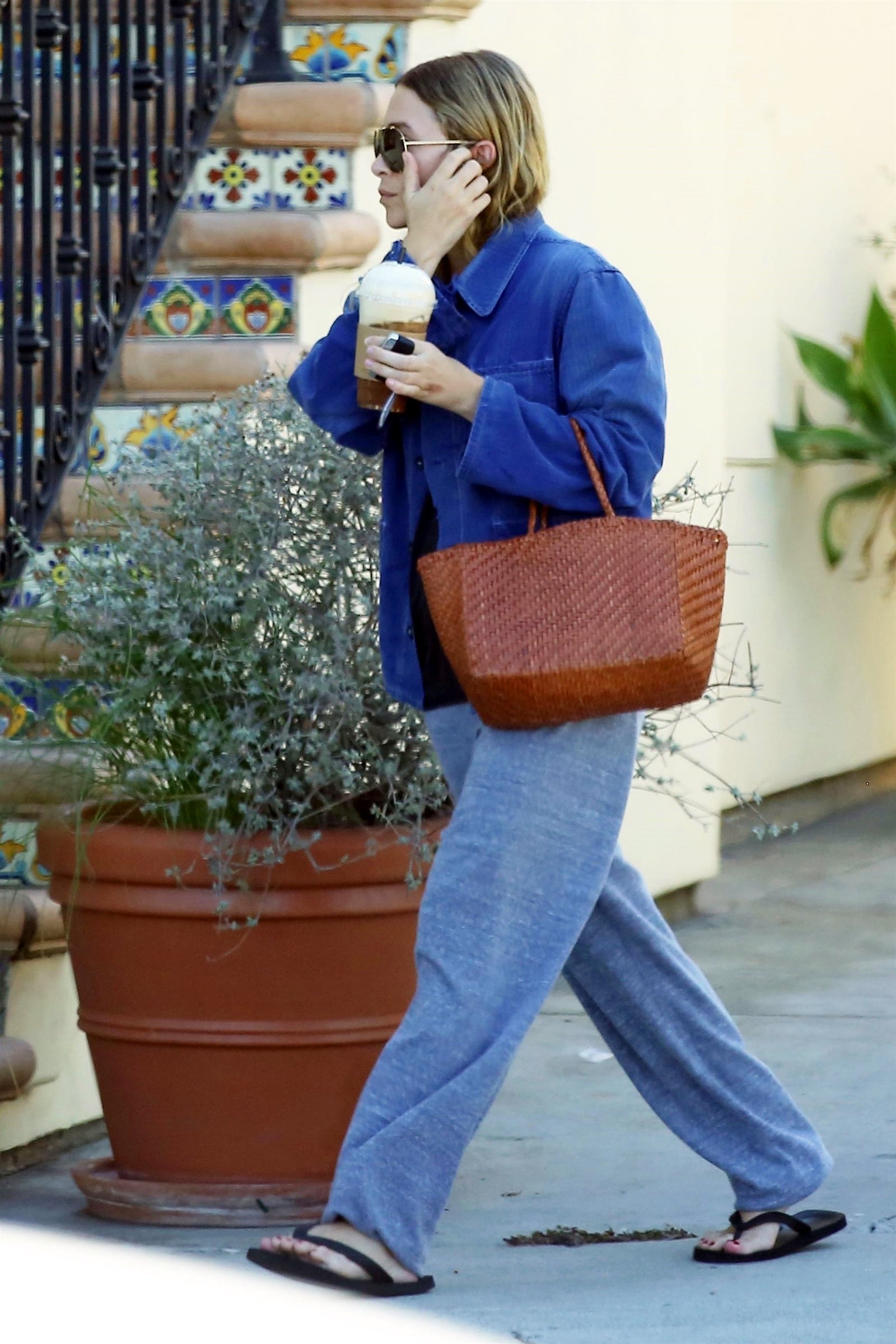 Ashley Olsen in Brentwood California carrying a coffee with wet hair, aviator sunglasses, blue canvas chore coat, camel brown Dragon Diffusion tote bag, baggy gray sweatpants, black flip-flop sandals