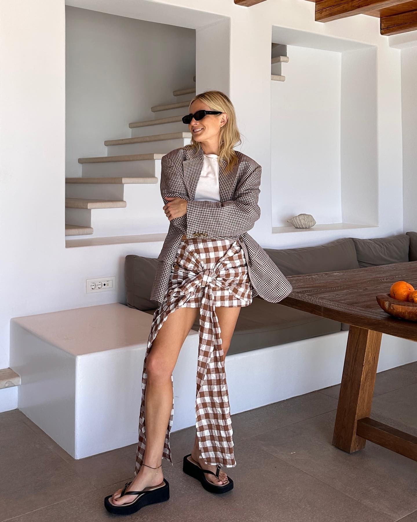 Fashion person Lucy Williams wears a gingham mini skirt with a tie front detail which falls past her left leg and right. She paired the skirt with 90-esque platform flip flops and a white t-shirt and check blazer. She is also wearing val sunglasses. This image was taken on her Instagram recently
