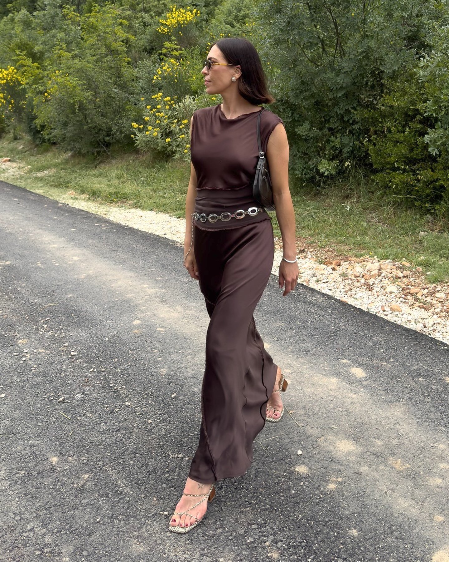 British influencer Jessica Skye wearing sunglasses, a small black shoulder bag, brown silky sleeveless dress, metal loop accent belt, and strappy neutral snakeskin heeled sandals