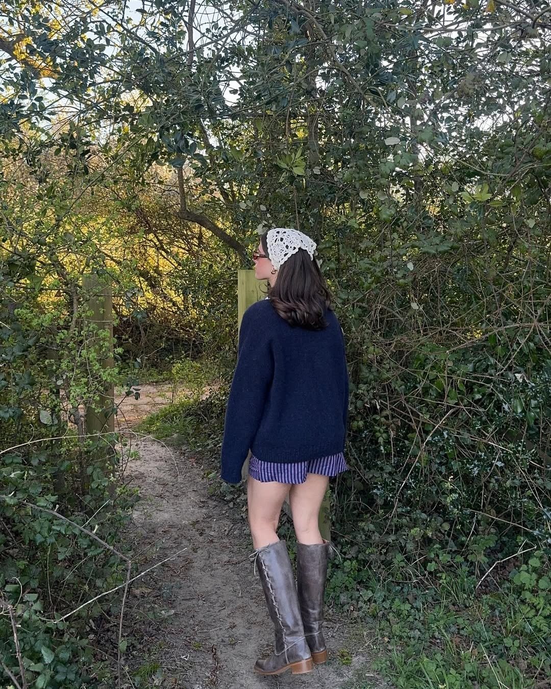 A woman standing wearing stripped boxer shorts, a blue sweater, and laced-up grey boots.
