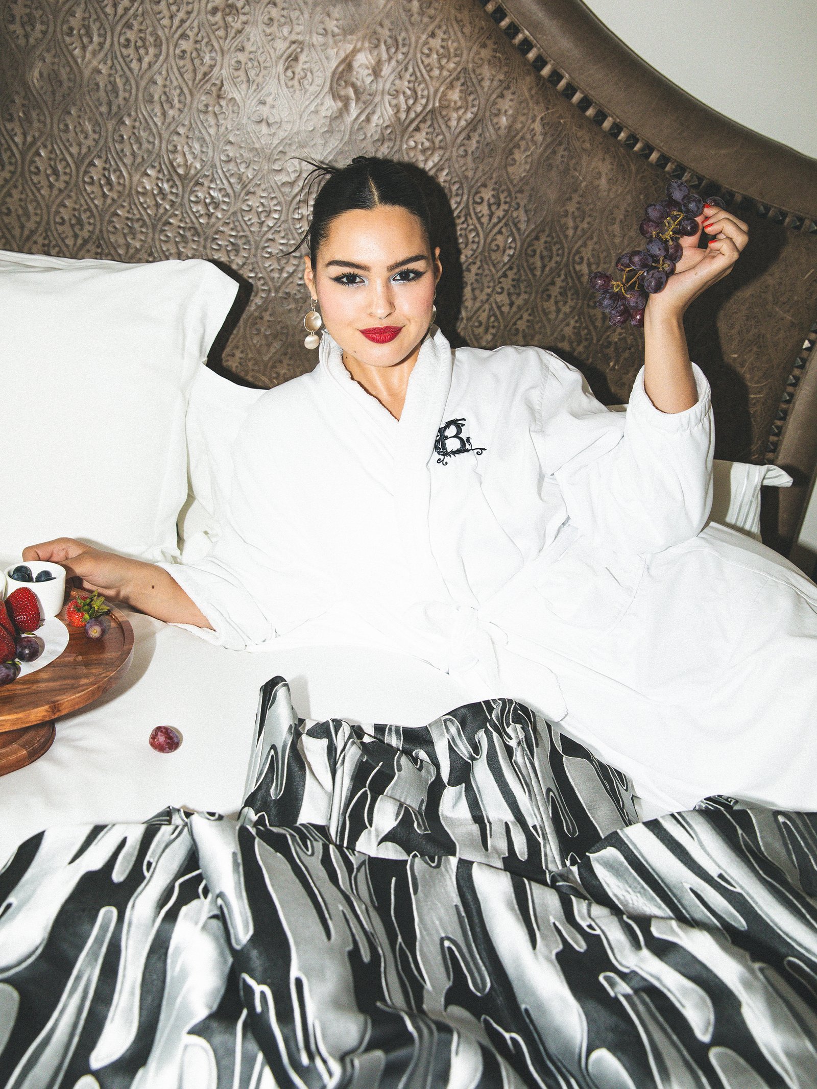 Anna Lambe is sitting in her hotel bed wearing a white robe and eating a fruit plate.