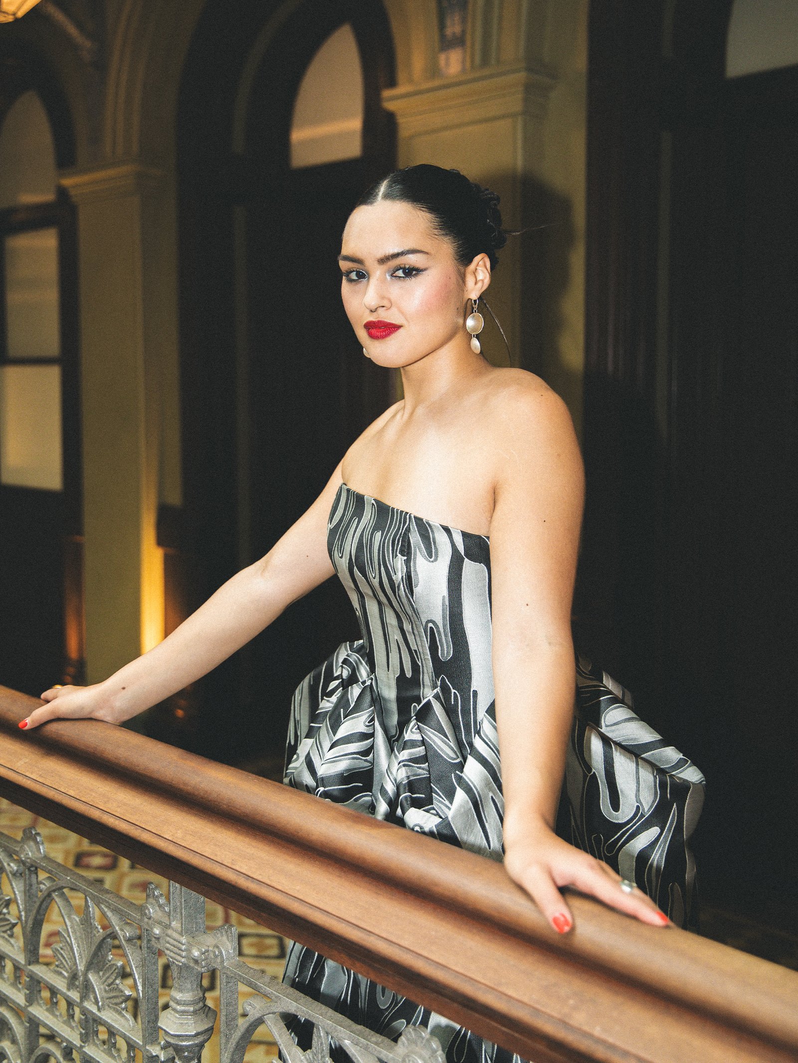 Anna Lambe stands against the railing of her hotel lobby. She is wearing a silver and black strapless gown with brocade design. Her hair is slicked back into a bun and she has a red lip.