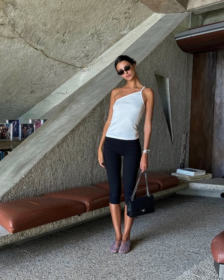 A woman standing wearing black capris, a white one-shoulder top, and clear jelly sandals.