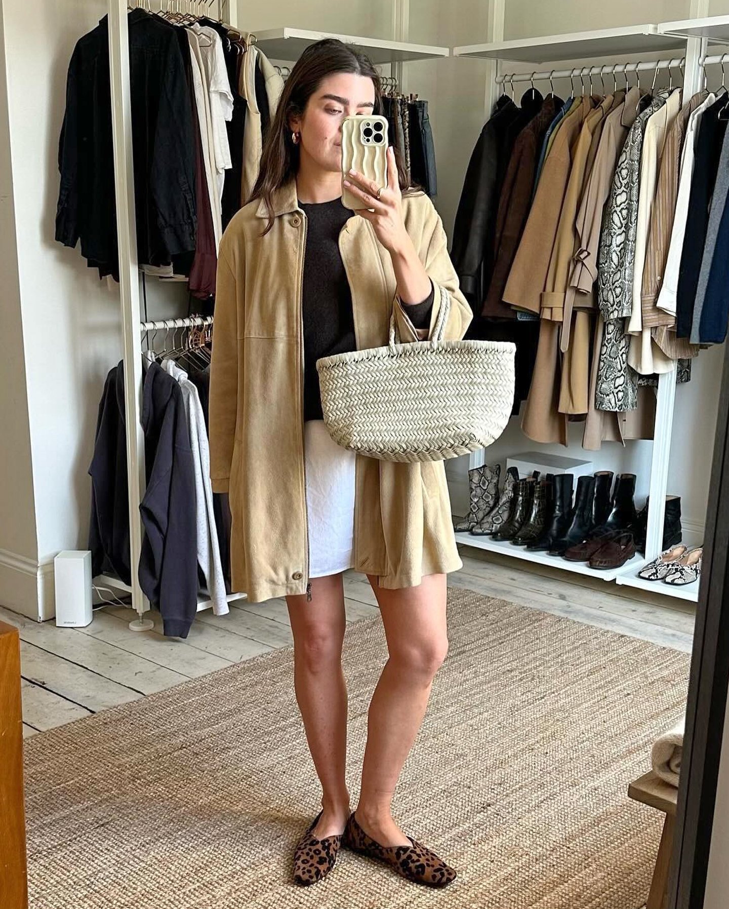 British style influencer Anna Newton poses for a mirror selfie in the closet of her London home wearing a tan suede jacket, black top, white woven tote bag, a white linen skirt, and leopard-print flats.