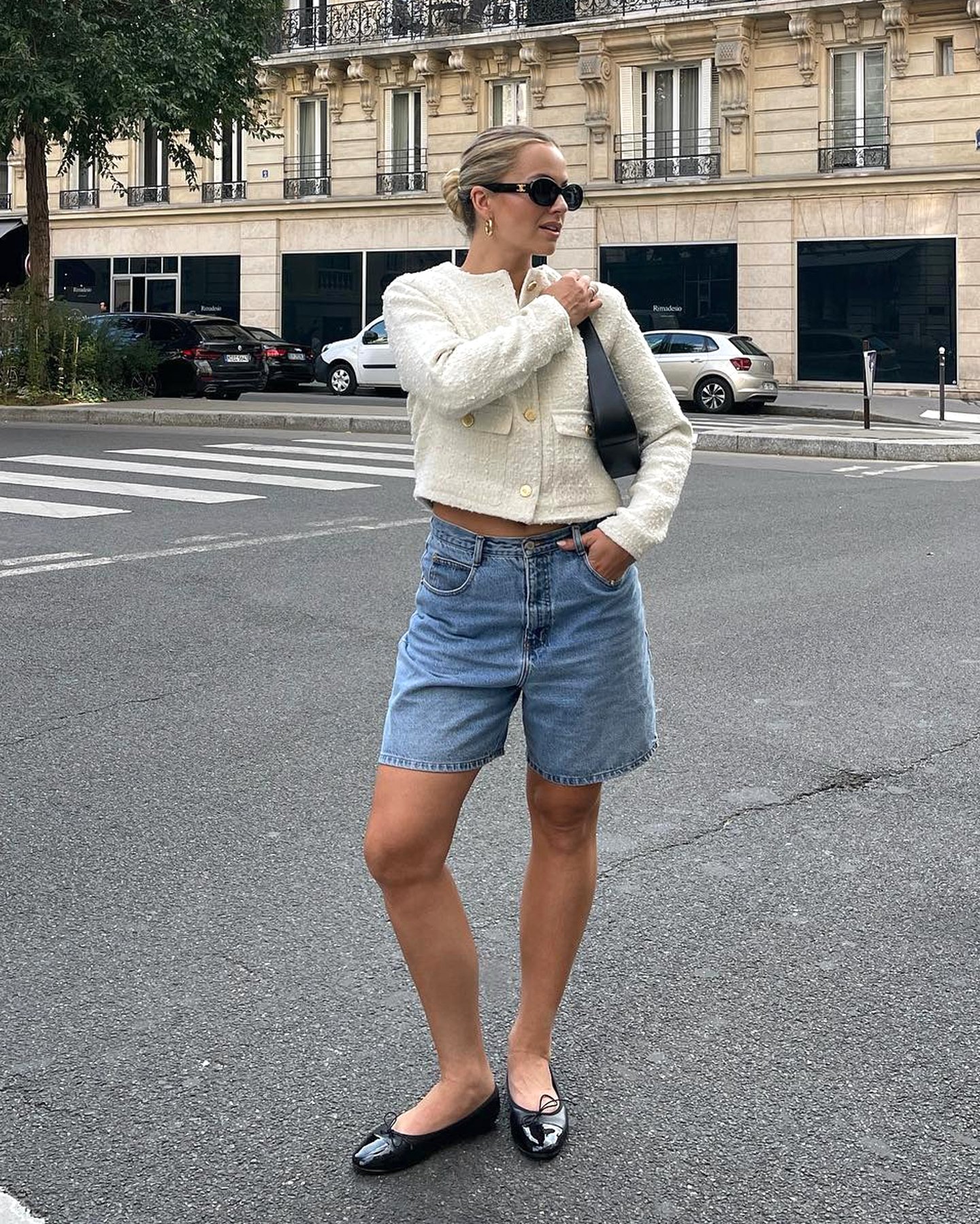 Style influencer Izzy Dilger poses on a street in Paris wearing black oval sunglasses, a cream tweed jacket, black mini shoulder bag, long denim shorts, and black patent ballet flats.