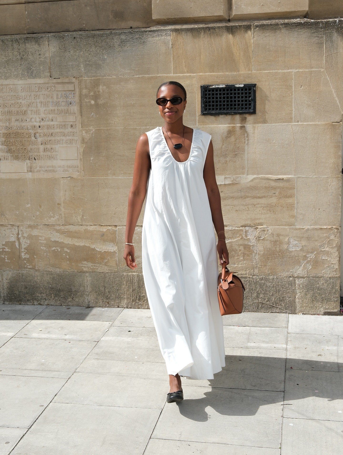 British Taffy Msipa poses in black oval sunglasses, a black cord pendant necklace, ruched white sleeveless dress, tan Loewe puzzle bag, and black ballet flats