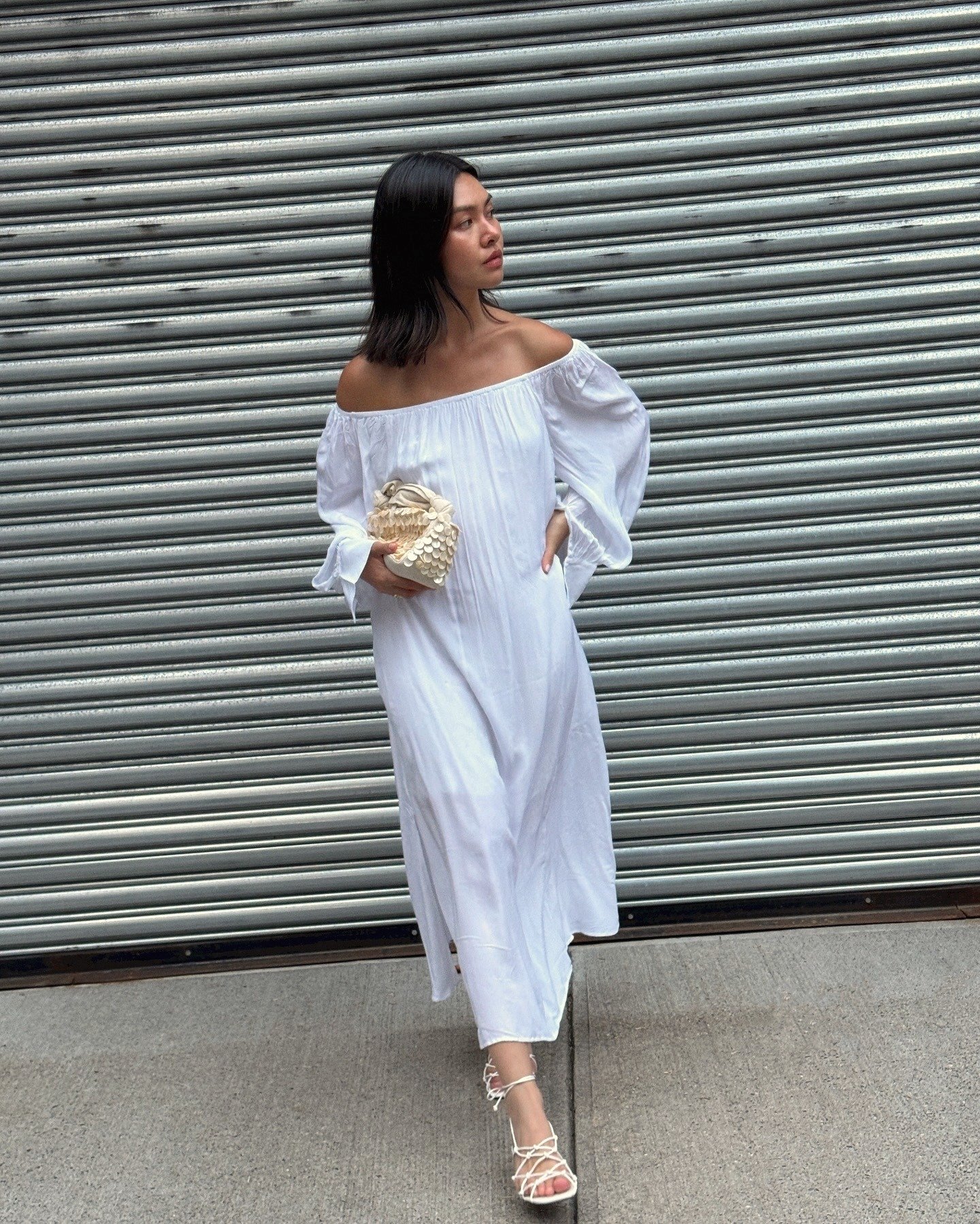 style influencer Mimi Nguyen poses on a NYC sidewalk wearing a white crinkled off-the-shoulder midi dress with balloon sleeves, a shell embellished clutch bag, and strappy white heeled sandals
