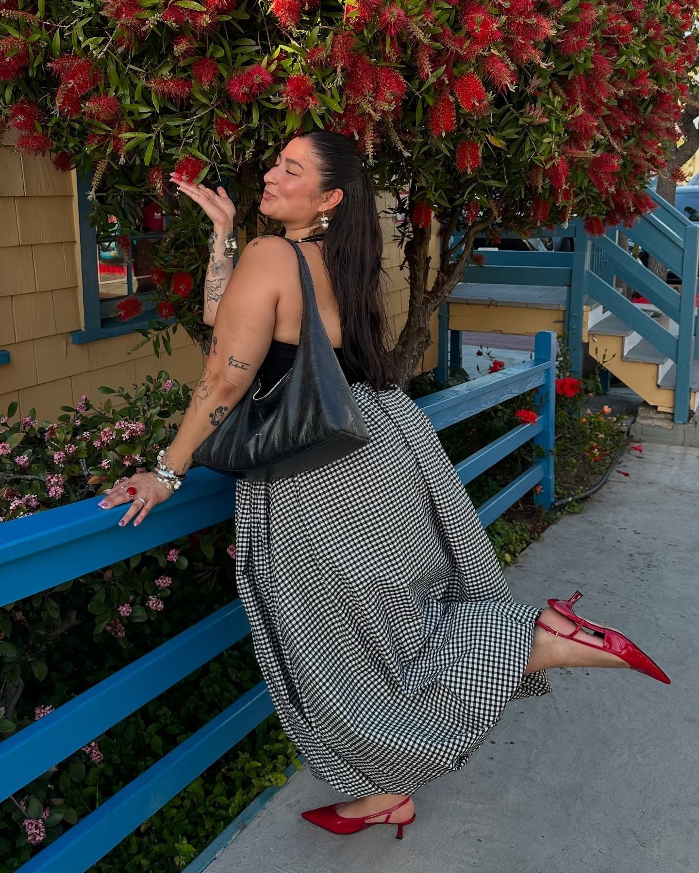 Fashion person Javiera wears a black and white gingham skirt, a black halter neck top read kitten heels and carried a black slouch shoulder bag. She is stood in front red flowers and is posing kissing one in a recent image taken from her Instagram.