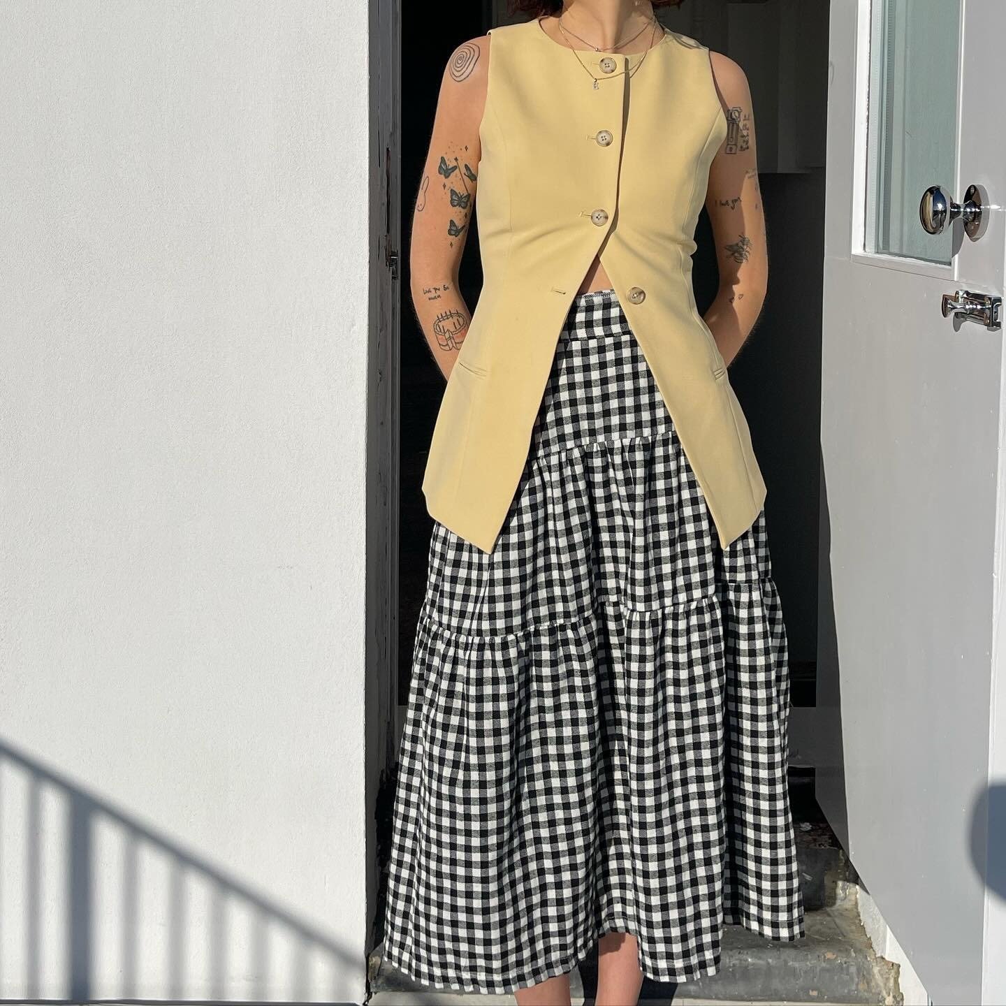 Fashion person Evie wears a butted yellow waistcoat and a black and white drop waist gingham skirt. She is stood in front of a white open door with a white wall on the other side in a recent image from her Instagram.