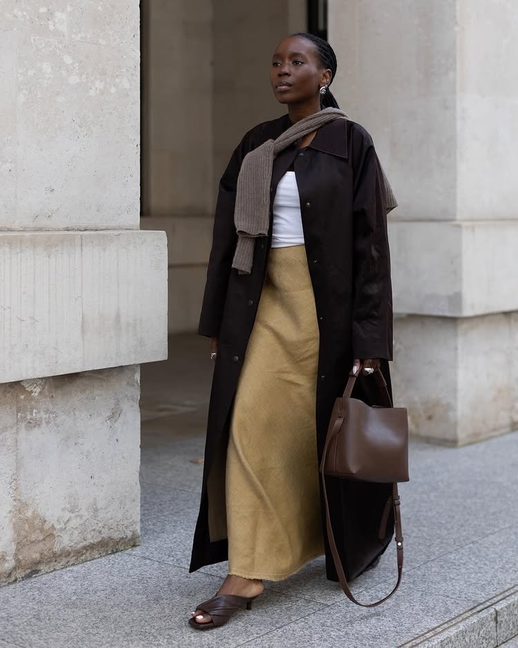 @daniellejinadu wears a pair of brown ruched sandals with a mustard skirt and black lightweight trench coat