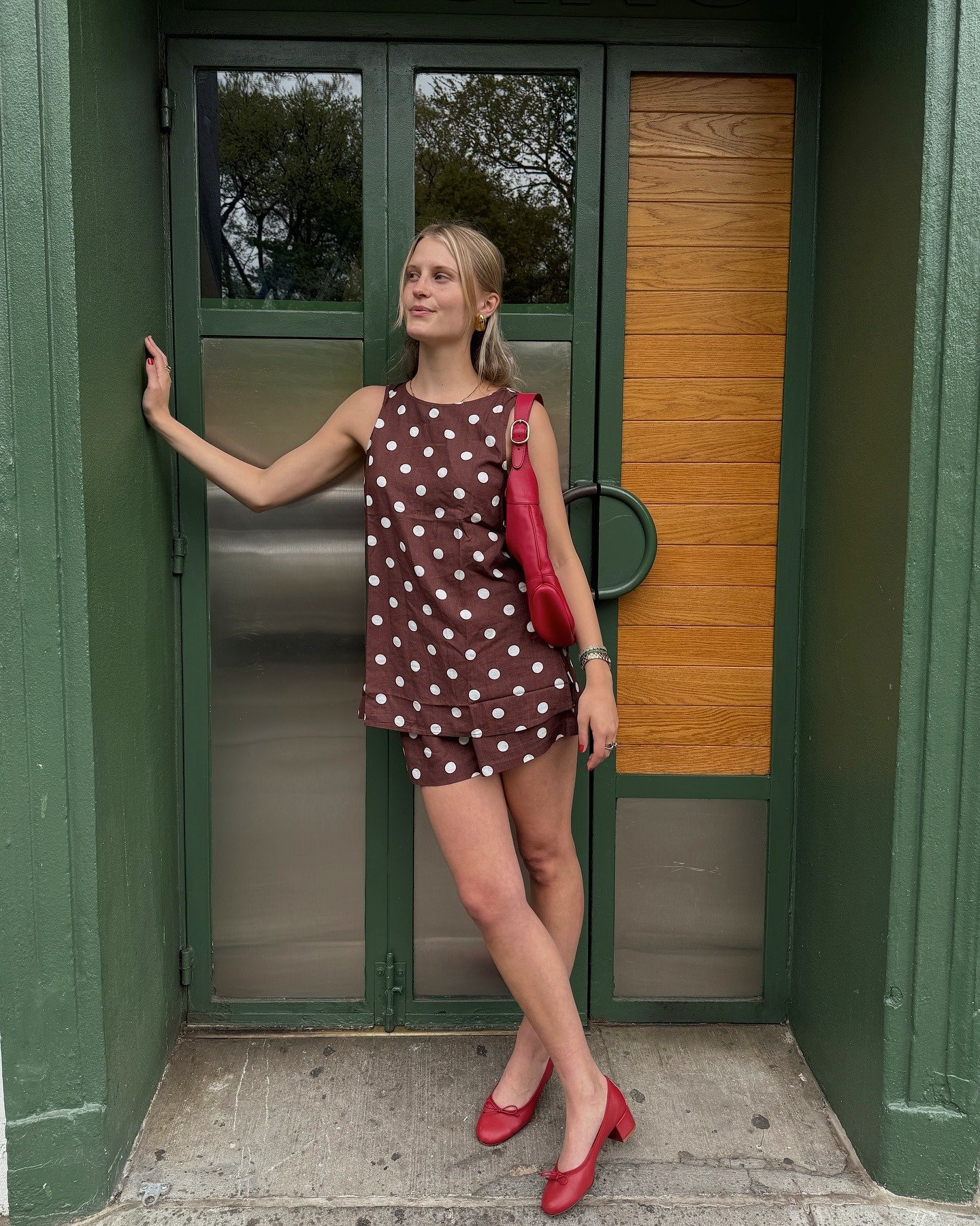 A woman in a brown and white polka dot set with a red slouchy bag and red ballet pumps.