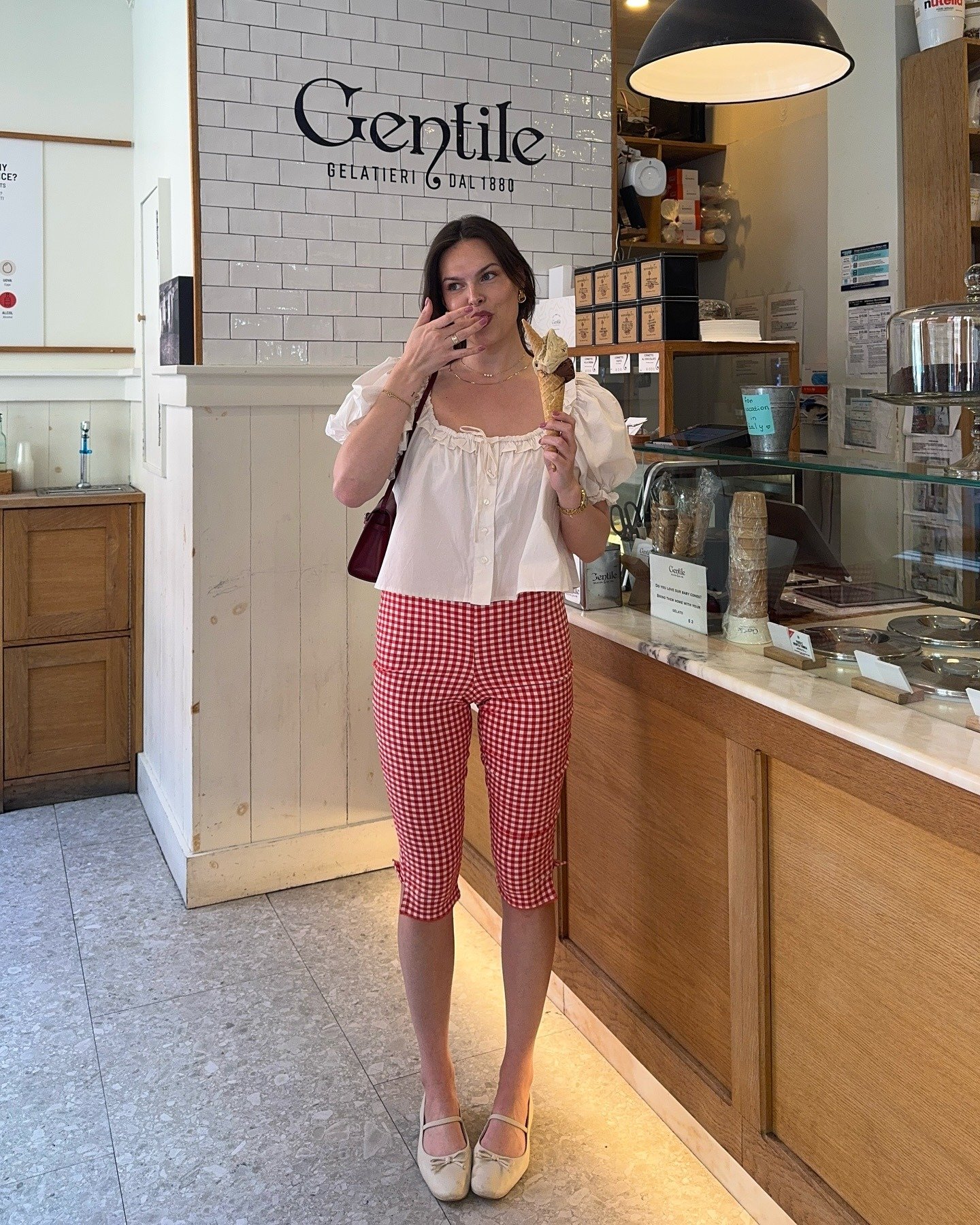 A woman wearing red and white gingham capris, a white ruffle blouse, and white ballet flats.
