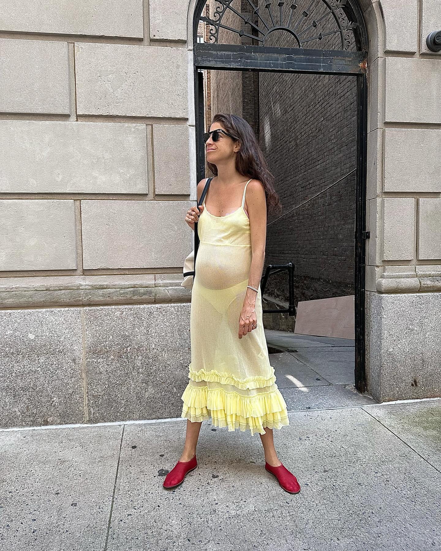 @leandramcohen wearing sheer yellow dress and red mesh ballet flats. She is stood in front of a brick wall in a recent photo taken from her Instagram.