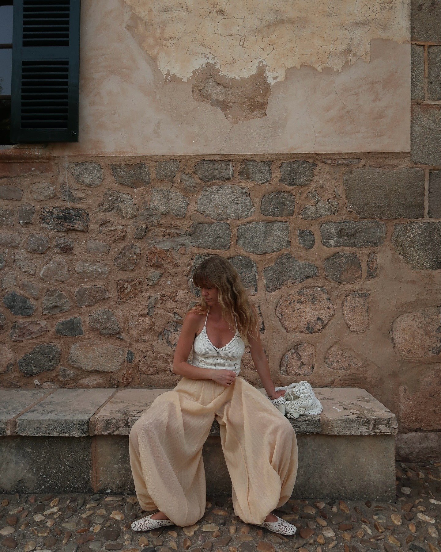 Fashion person Jeannette wears a crochet halterneck top, beige genie pants and crochet mules. She is sitting on a stone bench, in front of a stone wall, in a recent image from her Instagram.
