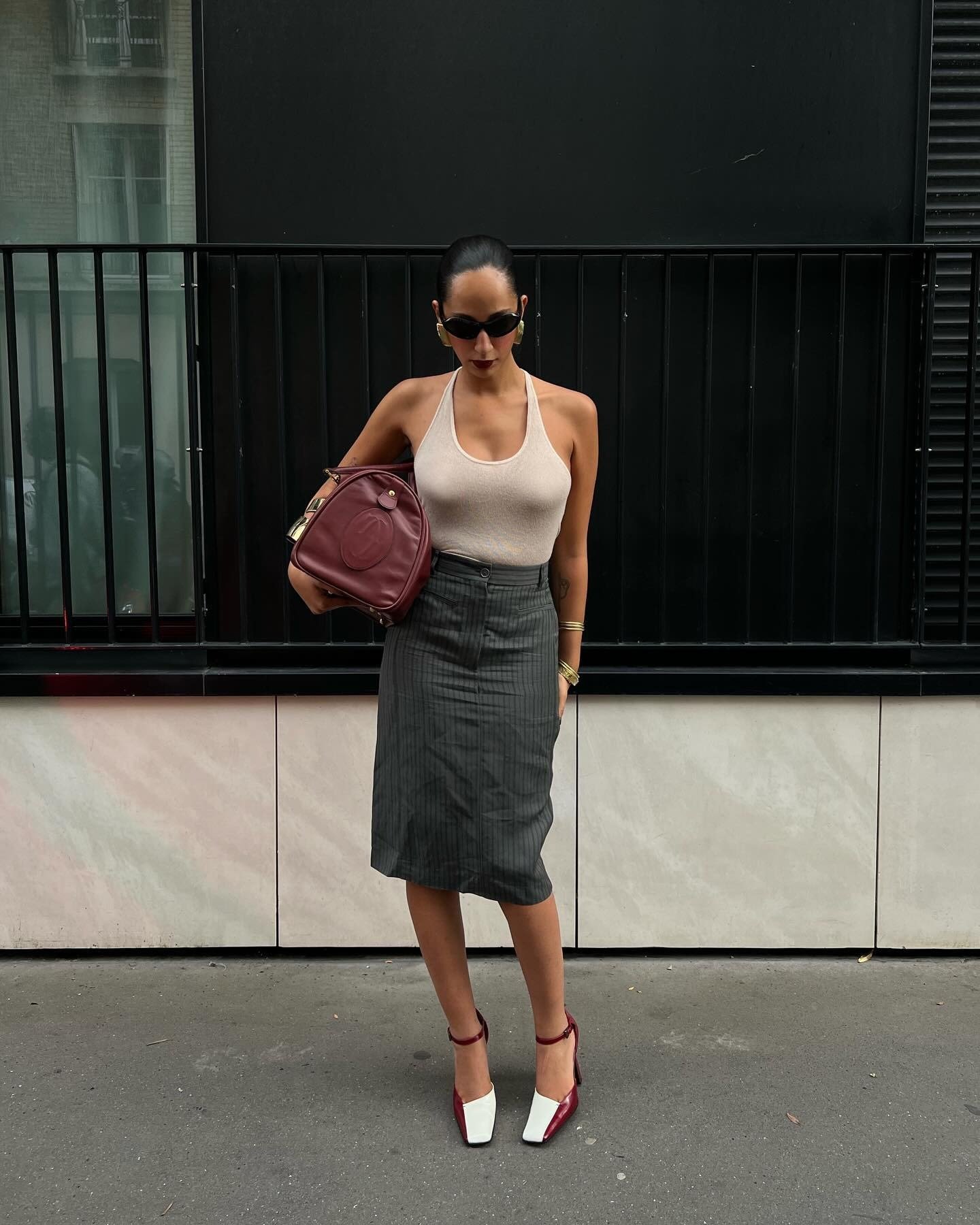Fashion person Emily Nitta wears a cream halterneck top, a tailored skirt and two tone heel. She is carrying a matching burgundy duffle and oval sunglasses in a recent image from her Instagram.