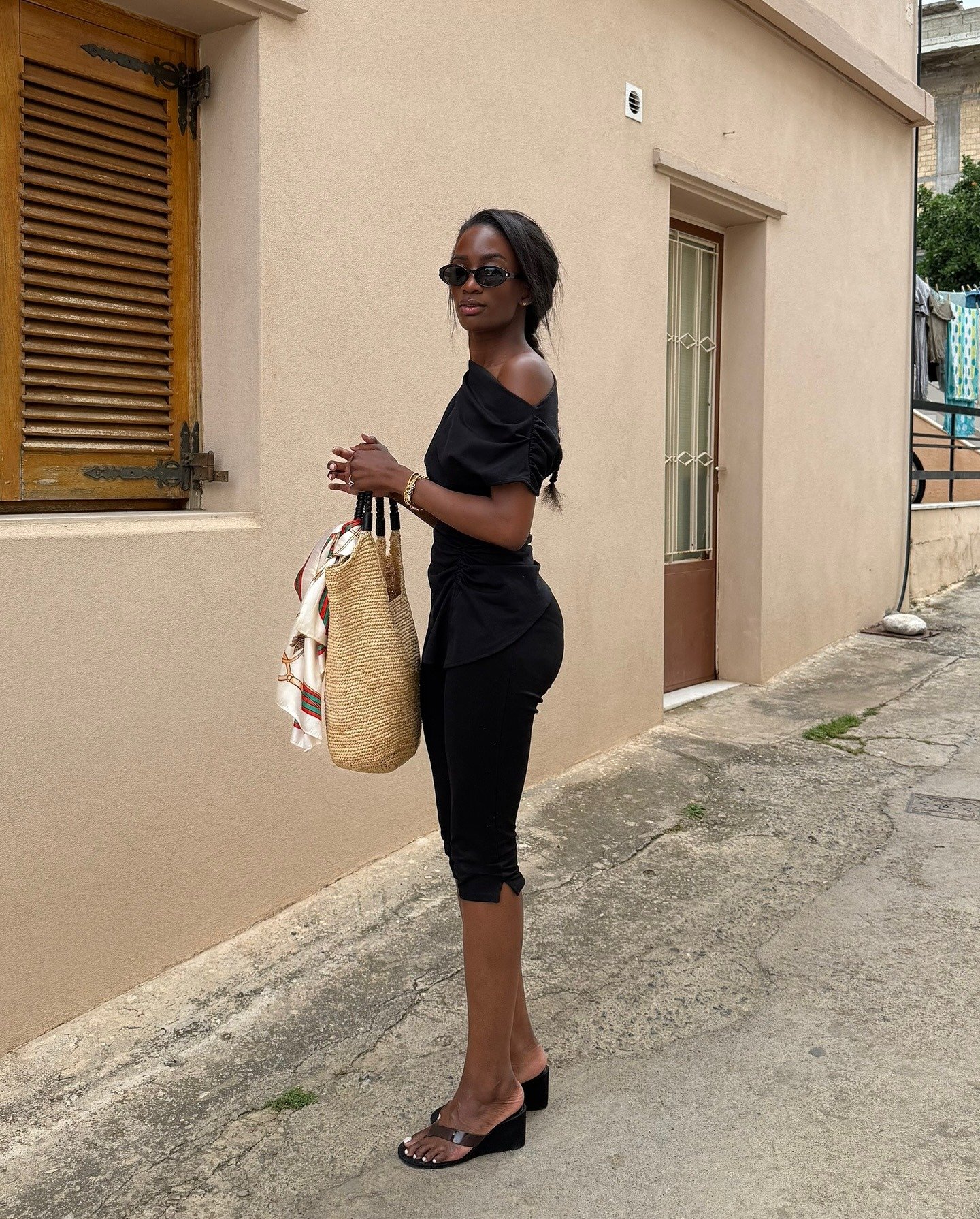 Fashion person Liv Madeline wears a black of the shoulder top, black carpis and black thong sandals and is carrying a crochet tote bag. She is stood in the middle of an old street on a trip away in a recent image from her Instagram.
