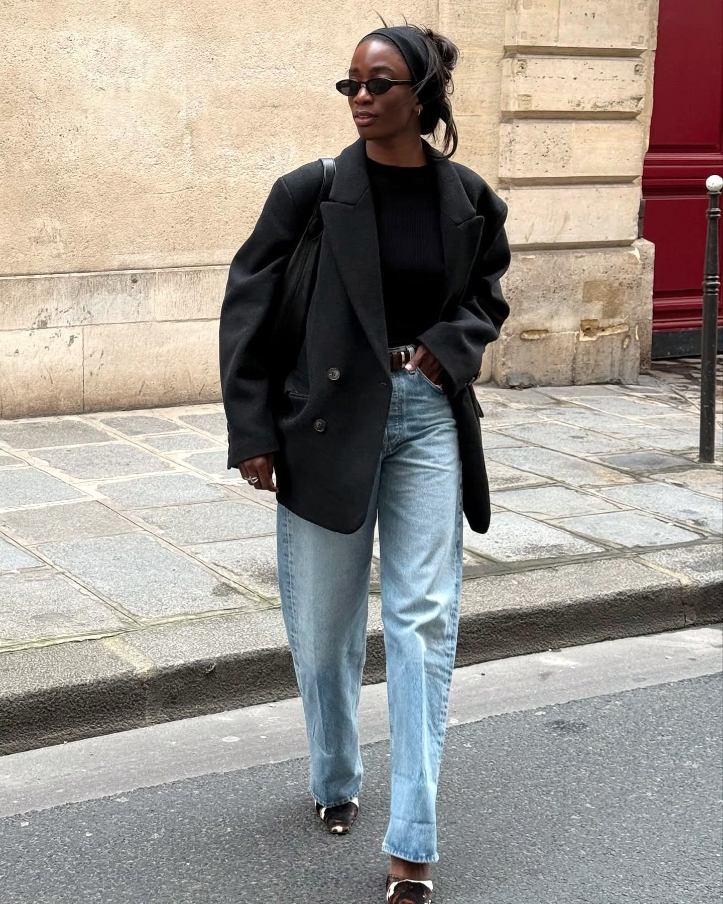 style influencer Liv Madeline crosses a street in Paris, France with a red door in the background wearing black oval sunglasses, a black head band, oversized black blazer, black top, belt, light-wash barrel jeans, and cow-print mule kitten-heels