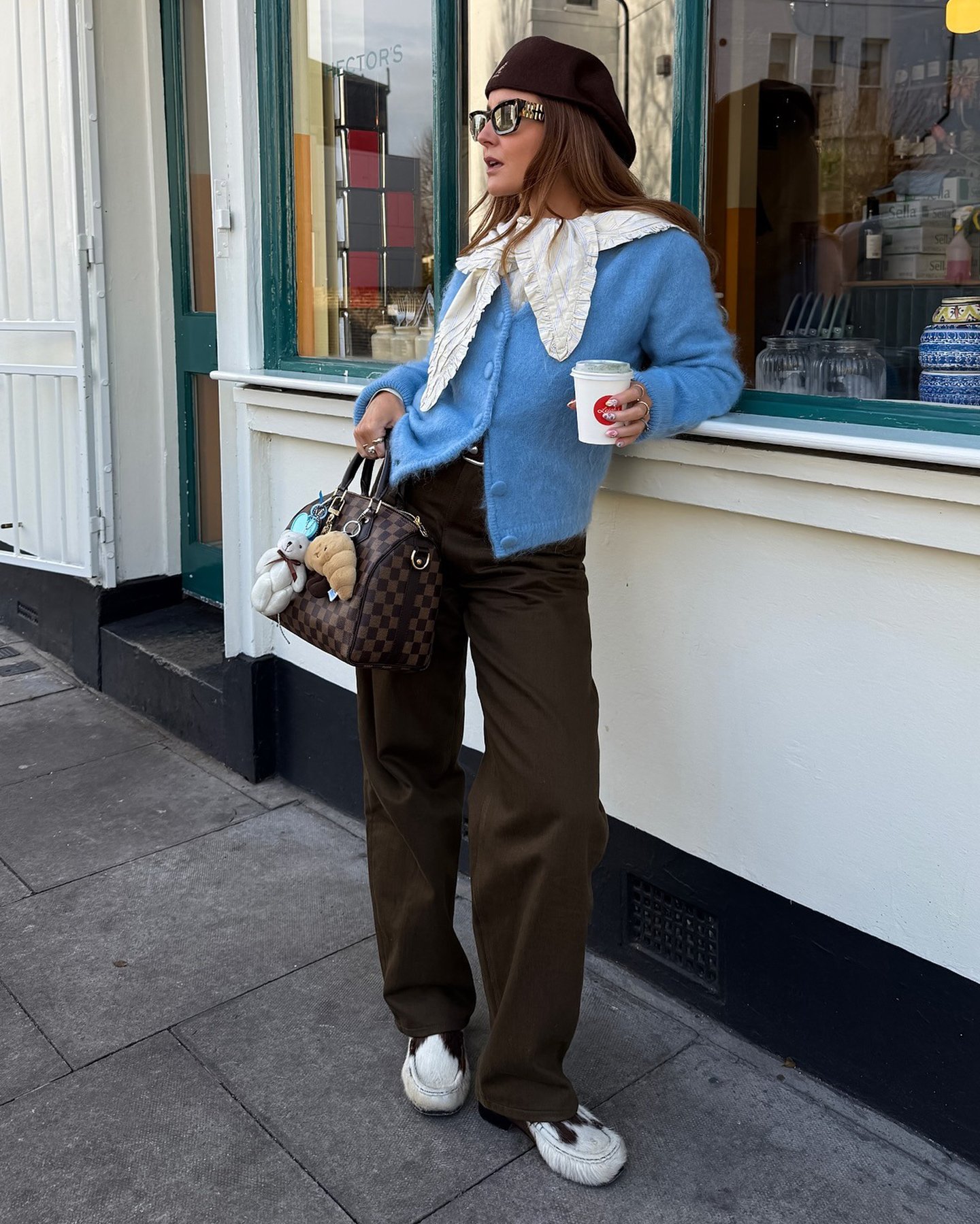 British style influencer Rebecca Ferraz Wyatt poses on a London sidewalk holding a coffee wearing a brown Kangol hat, ruffle collar, light blue cardigan sweater, a Louis Vuitton bag with bag charms, brown jeans, and cow-print loafers
