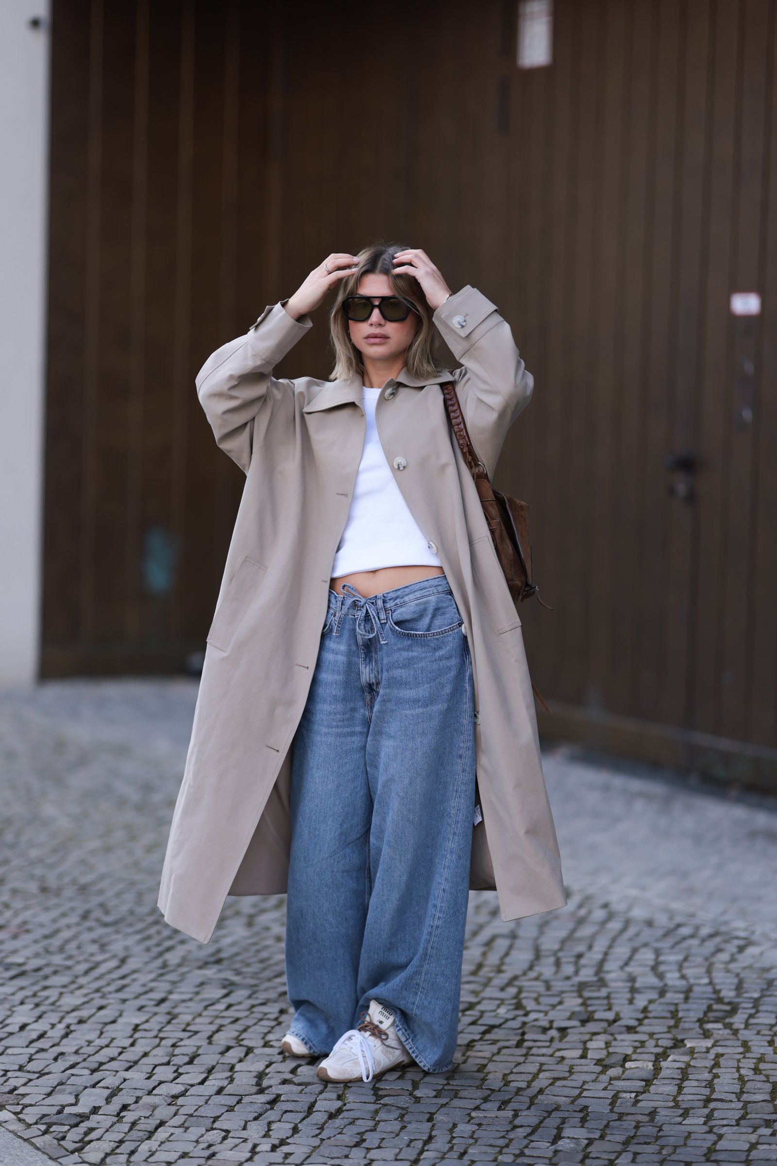Fashion Person Sophia Geiss at Berlin fashion week. She wears the drawstring jeans trend and pairs them with a brown trench coat, white Miu Miu x New Balance trainers a white cropped tee and black sunglasses.