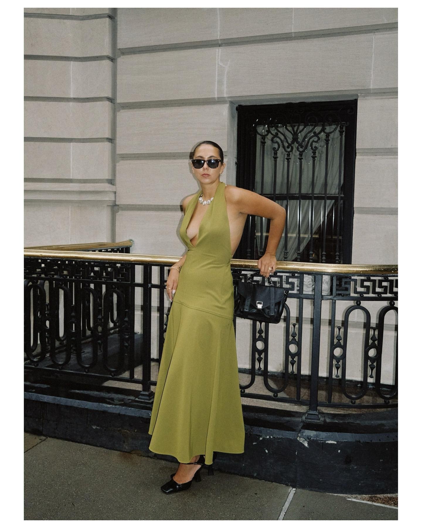 woman wears green halterneck maxi dress with black accessories leaning against a railing.