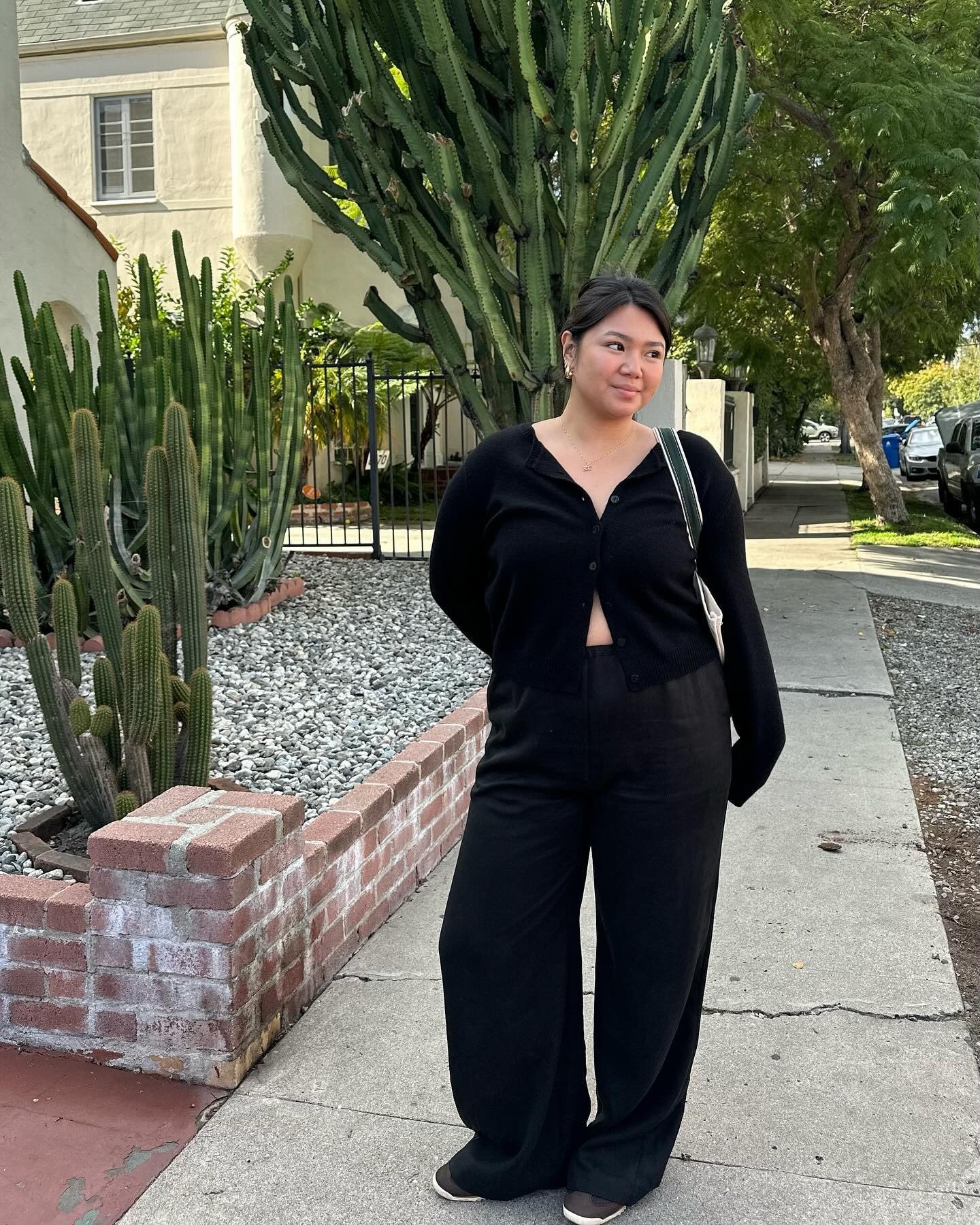 Influencer @marina_torres wears black satin trousers with black trainers and a partially unbuttoned black cardigan.