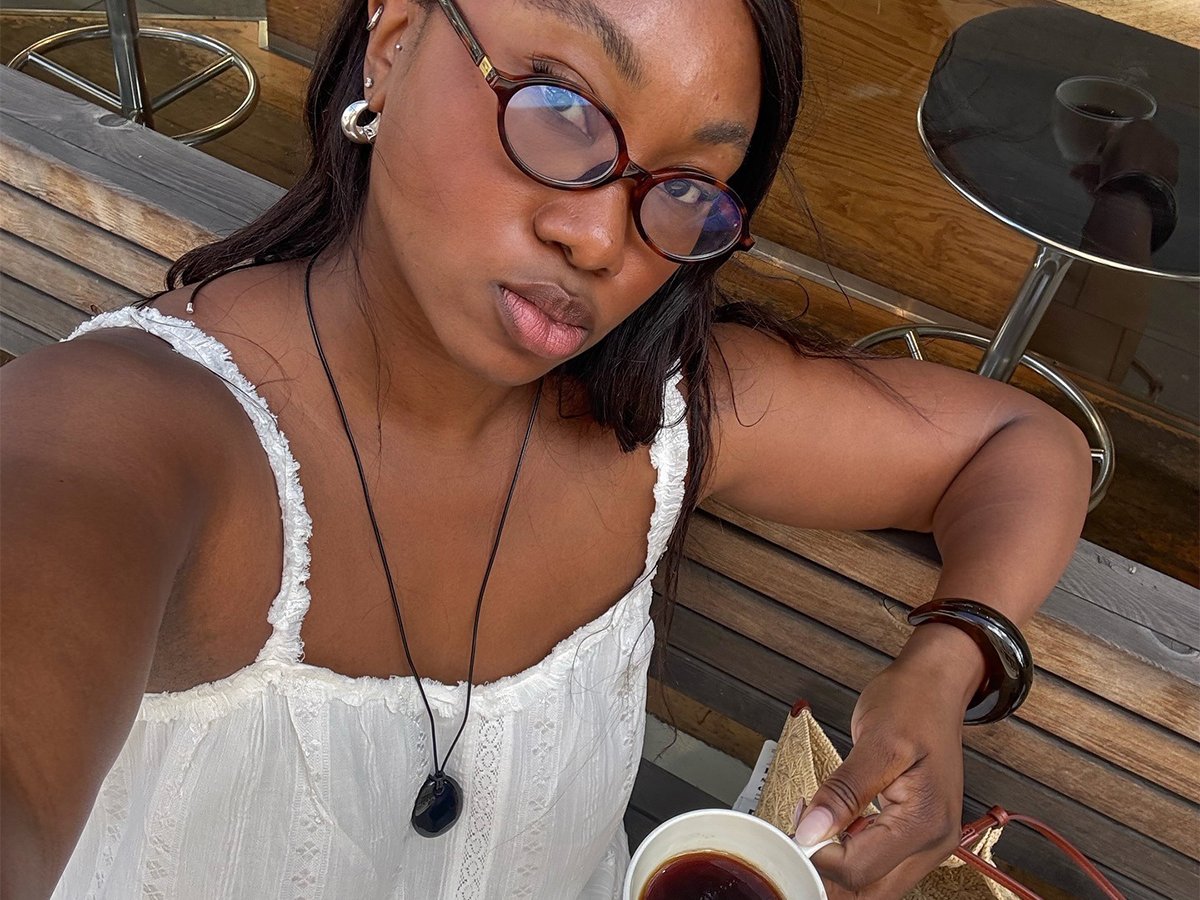 Influencer Nnenna wears a white lace dress and a long pendant necklace while holding a cup of coffee.