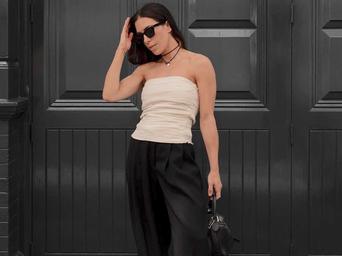 influencer Jessica Skye poses in front of a black wall in London wearing black sunglasses, a black cord necklace with a silver pendant, a beige ruched sleeveless top, black trousers, and a black bag