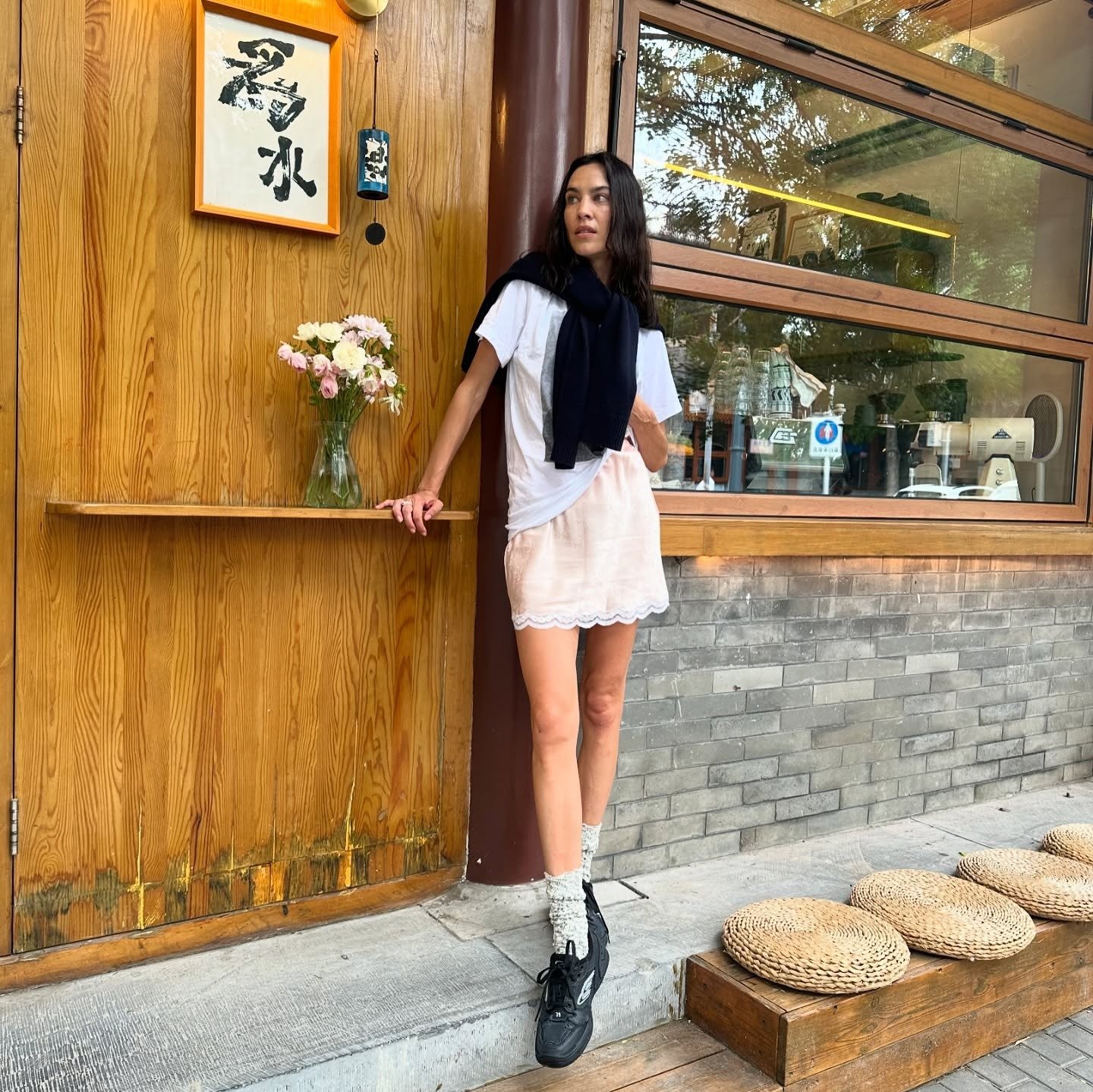 Alexa Chung stands outside wearing a pink satin miniskirt with lace-trim detailing with black trainers, a white t-shirt and a jumper wrapped around her shoulders.