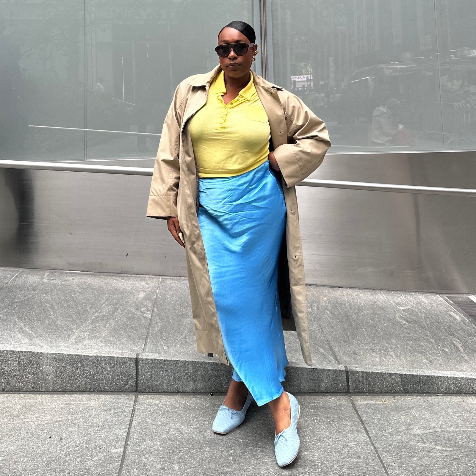 Editor Aniyah Morinia in blue satin skirt and yellow polo top with trench coat and blue ballet flats.