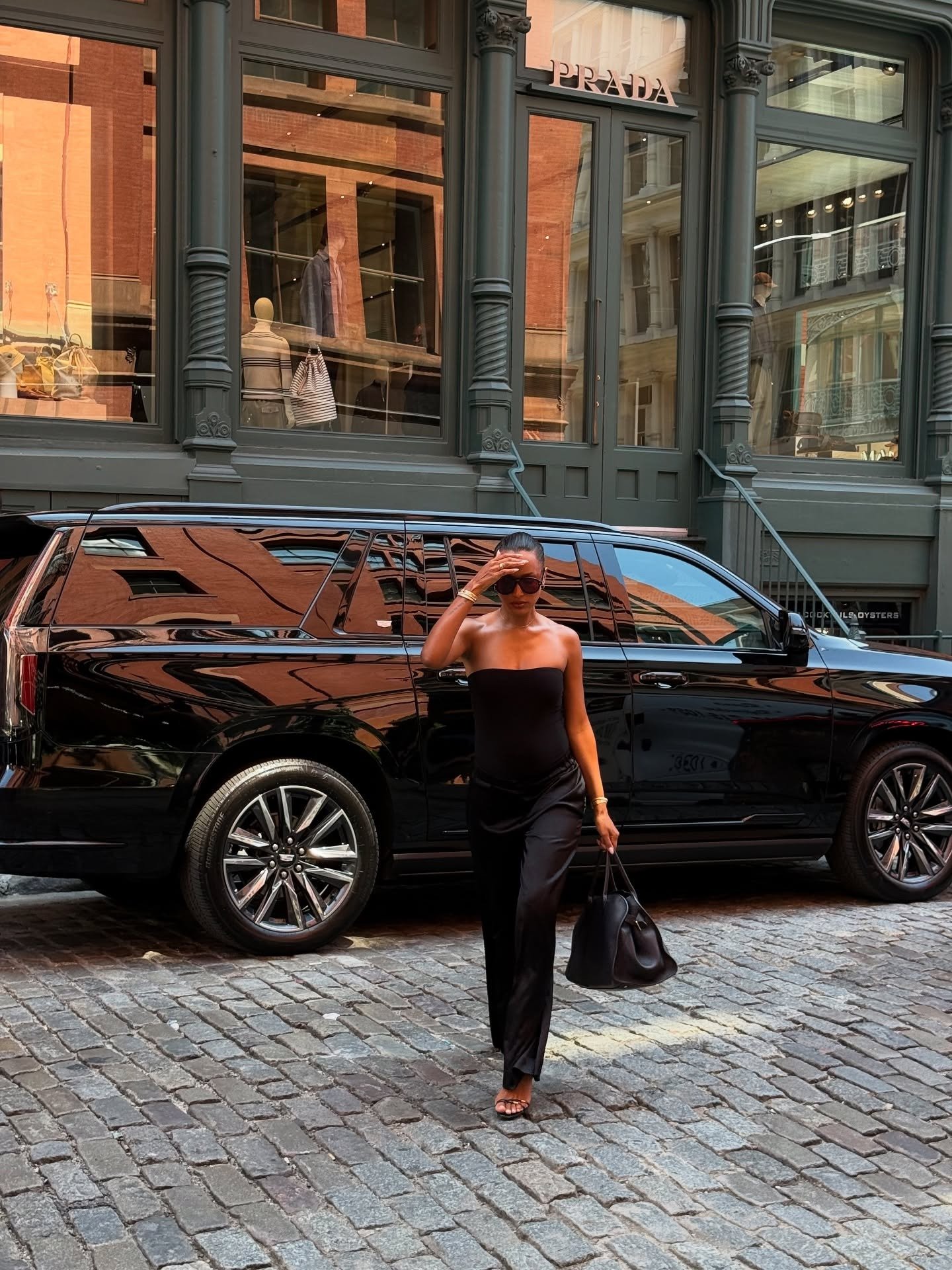 Model Jasmine Tookes wears black satin trousers with a black strapless top and black The Row Margaux bag.