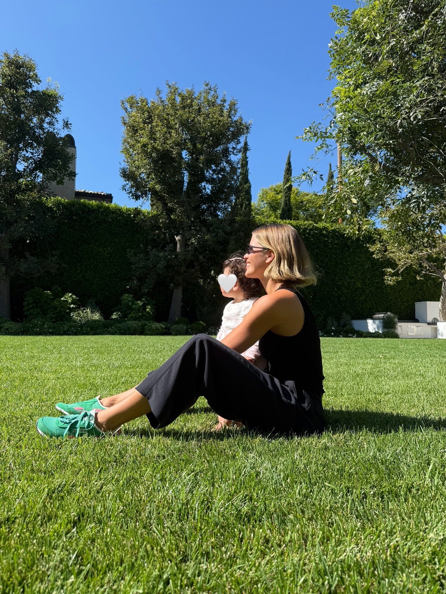 Sofia Richie sits outfit on the grass wearing black trousers with a black top and green trainers.