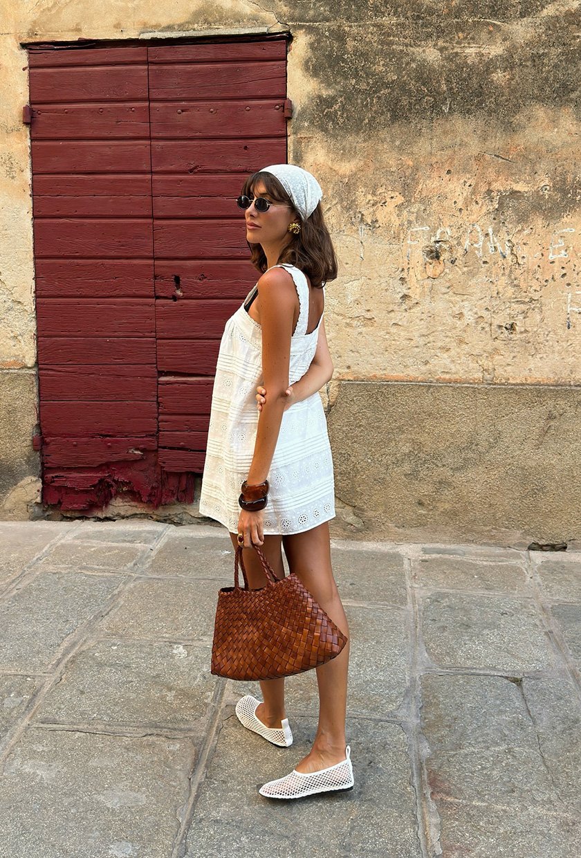The summer aesthetic, coastal boho is shown in a street style photo of a woman standing on the sidewalk wearing a white headscarf with black sunglasses, gold earrings, a white eyelet mini shift dress, stacked brown bangles, a brown woven leather tote bag, and white mesh ballet flats