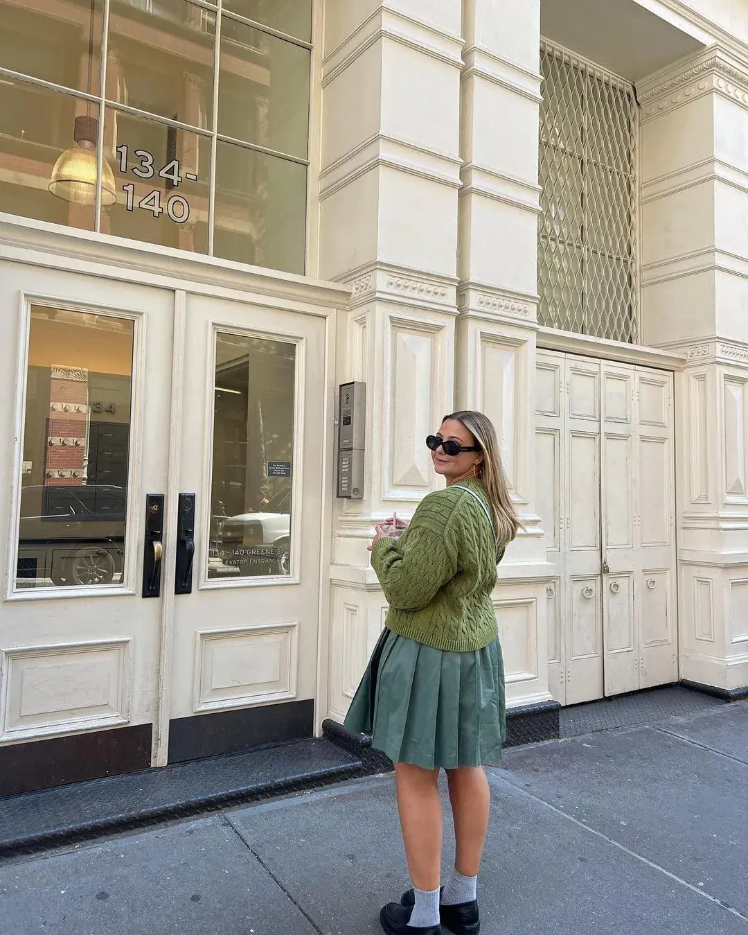 Influencer @oliviamarcus wears a dill green cardigan with a darker green pleated skirt, black sunglasses and glossy black leather loafers.