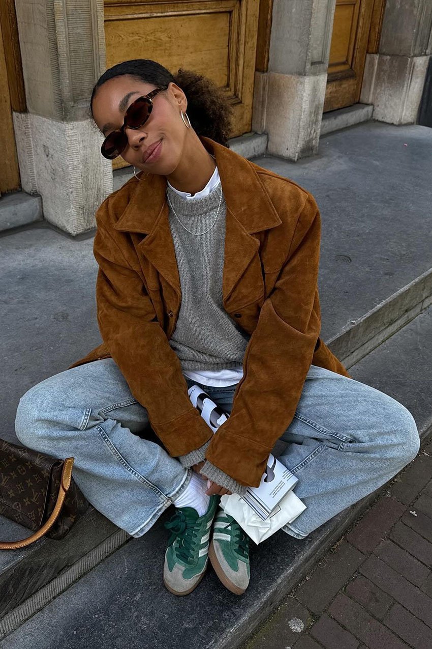 A woman wearing a brown suede jacket, gray sweater, blue jeans, and green suede sneakers